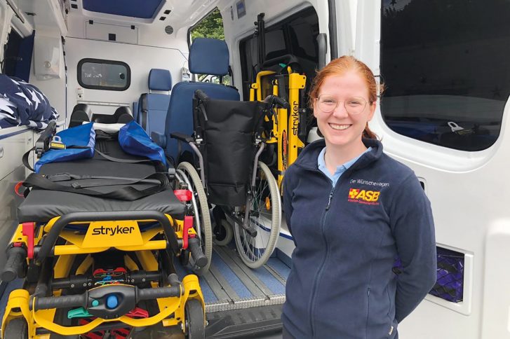 Eine junge Frau steht vor einem Krankentransport. Sie lächelt. Der Wagen ist offen. Mansieht die Krankliege, einen Rolllstuhl und das innere des Wagens.