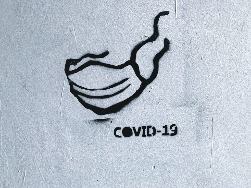 Grafitti einer medizinischen Maske auf einer Mauer. Darunter steht: Covid-19 – Isolation Quarantäne – Eindämmungsverordnung