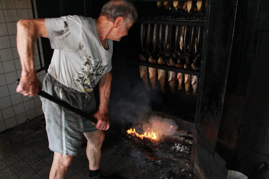 Hans Werner Schwan schippt Holzspäne in den Ofen. Noch züngelt die Flamme. Es ist die erste, heißere Garstufe, bevor es zum reinen Räuchern geht.