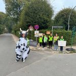 Cornelia Lewin lässt sich für die kuhle Verkehrsaktion das Fell pber die Ohren ziehen – im übertragenen Sinn, ist ja klar. Als Verkehrskuh will sie Öffentlichkeit für die Sicherheit der Kinder schaffen. Im Hintergrund sind die Schulleiterin Frau Appelt (in der Mitte), zwei Klimadetektive, ein 2. Klasse Schüler und vier weitere Elternratsmitglieder zu sehen. // Foto: privat Keyword:Lehmkuhlenweg Verkehrsaktion kuhle